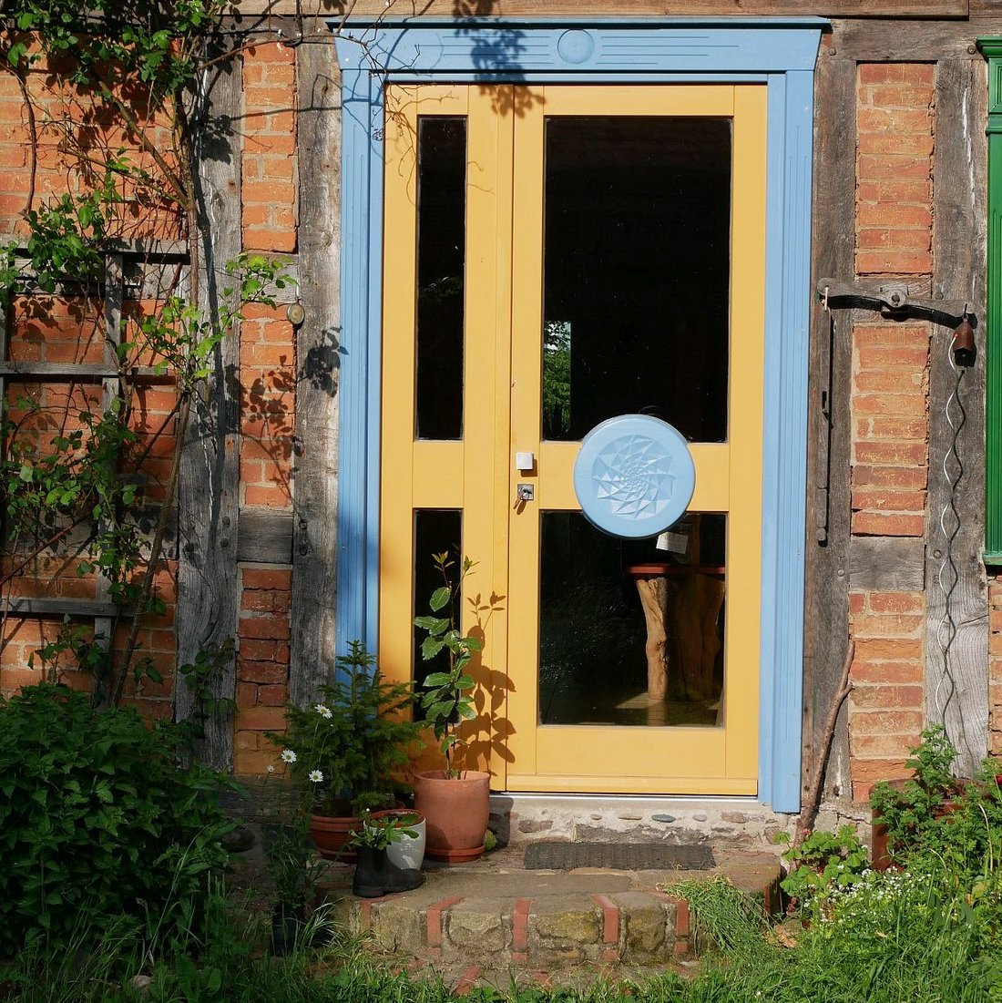 Eine einladende zweiflügelige Haustür in den Farben Hellblau und Gelborgange mit einem geschnitzen Ornament, ähnlichen einem Mandala) auf einem Türflügel. Die Türfüllungen sind aus Glas.