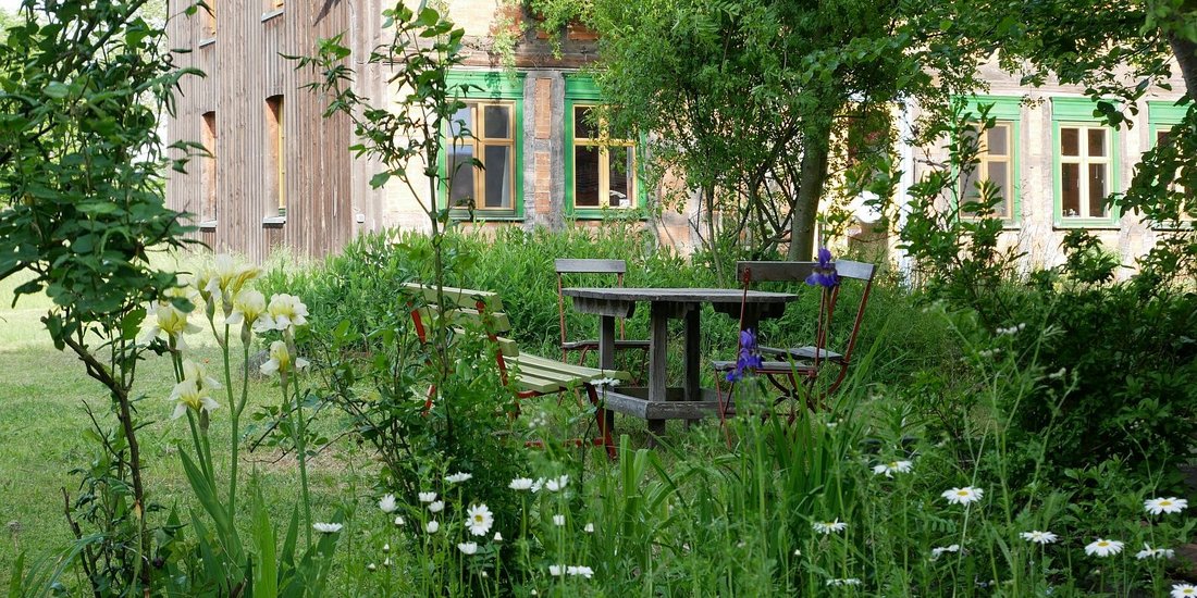 Ein Sitzplatz im Grünen, Tisch und Stühle auf dem Innenhof von Hof Erdenlicht, im Vordergrund eine Blumenrabatte mit bühenden Margariten und Lilien
