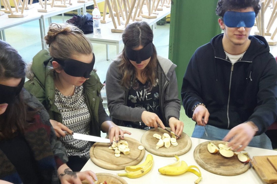 Jugendliche sitzen um einen Tisch, ihre Augen sind mit Schlafmasken abgedeckt. Sie schneiden Obst auf Scneidbrettern für einen Obstsalat.